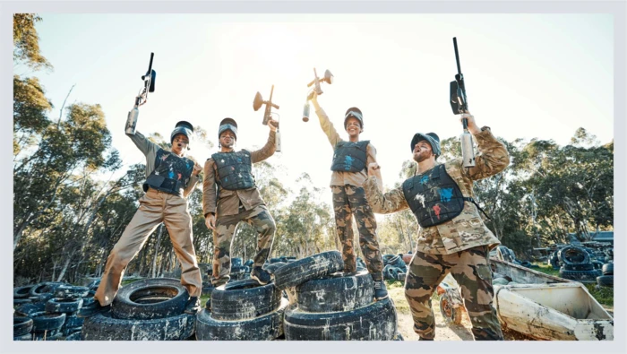 A group of friends dressed in paintballing gear hold their paintball guns in the air for a group photo.