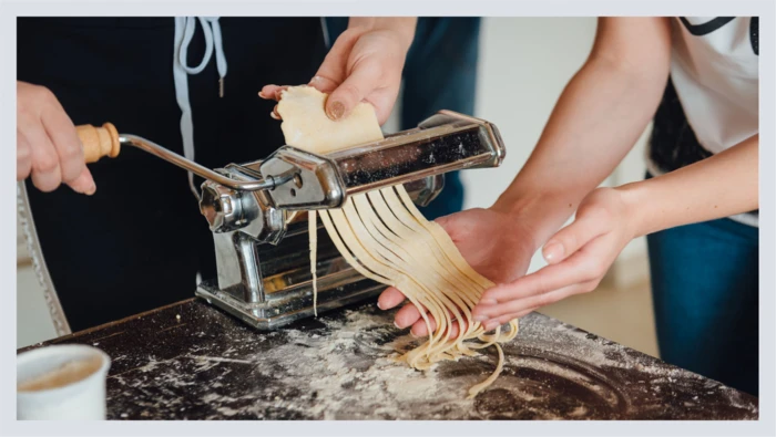 A closeup of a pasta-making machine shows pasta being made on a table doused with flour. 