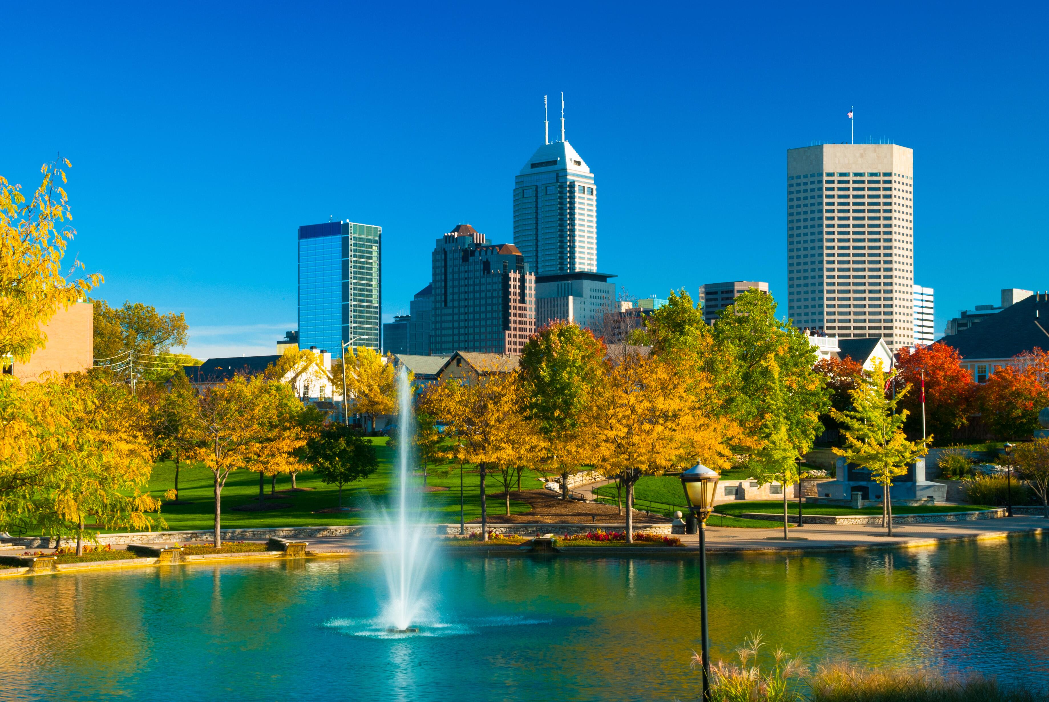 Downtown Indianapolis skyline with the Indiana Central Canal with a fountain, surrounded by green, yellow and red trees and park land, during Autumn