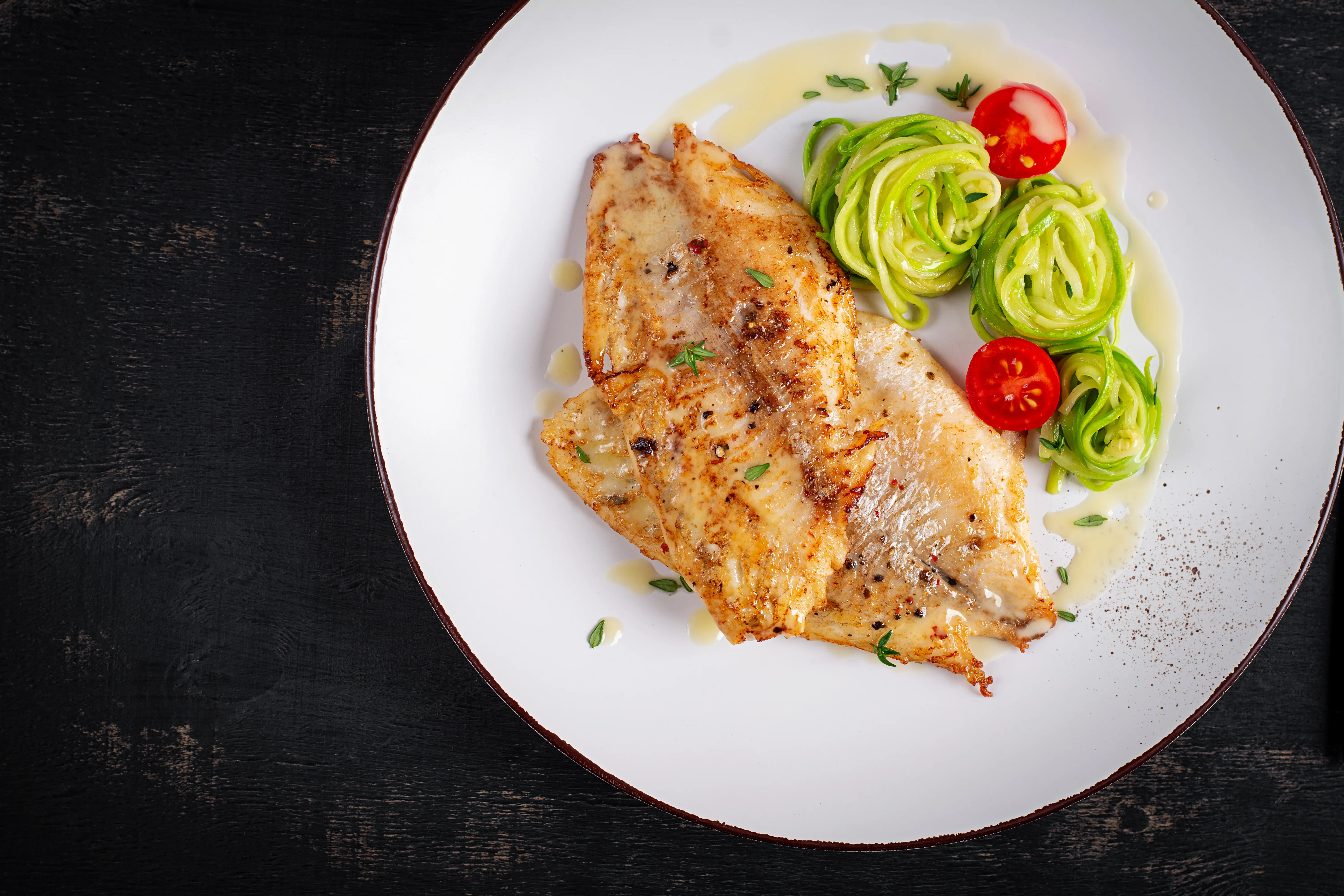 Top view of grilled fish fillet with zucchini pasta on a white plate, black table top