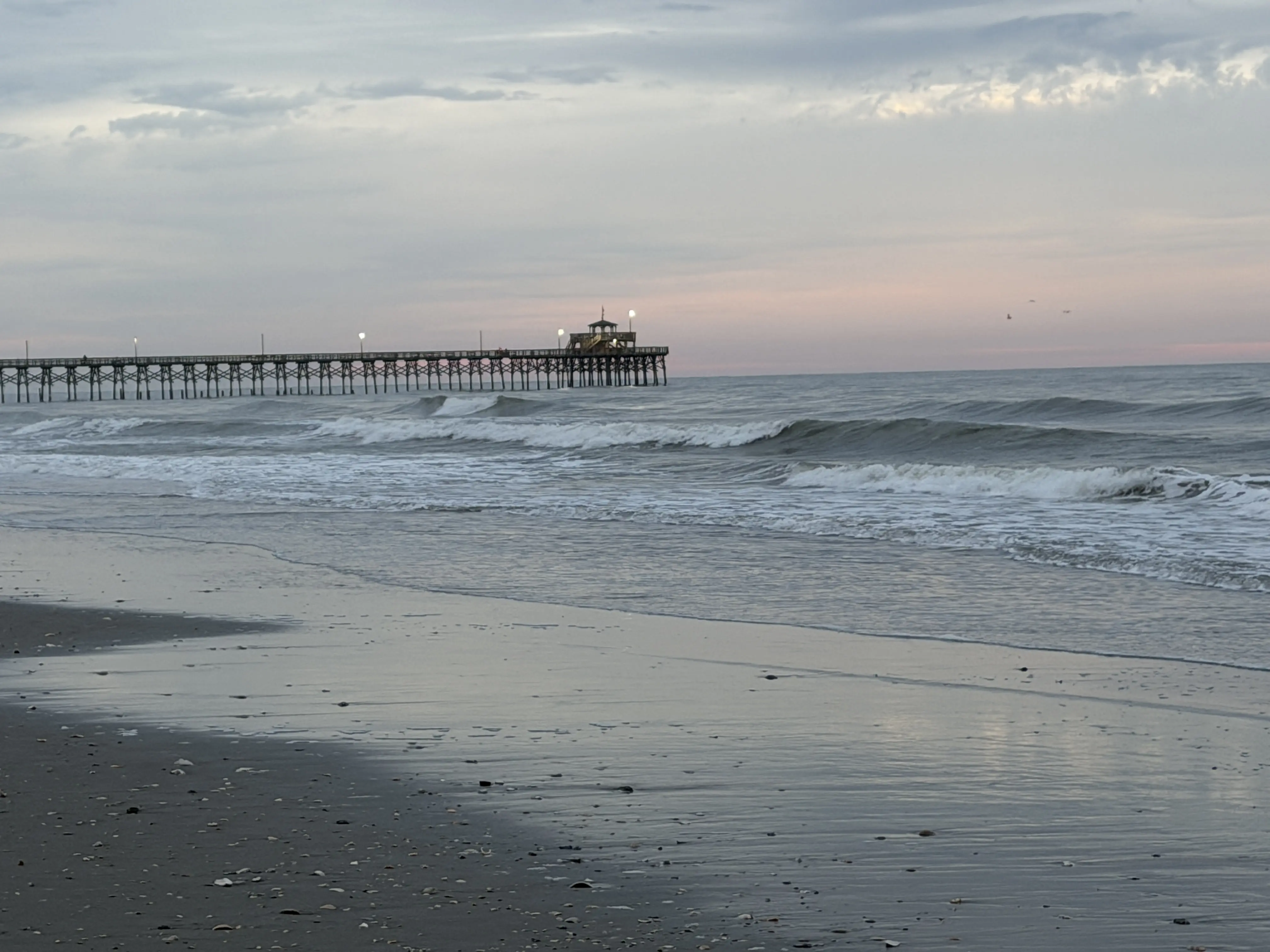 sunrise on dimly lit beach with a streak of pink sky at the horizon. A pier the reaches out into the ocean sit in the background