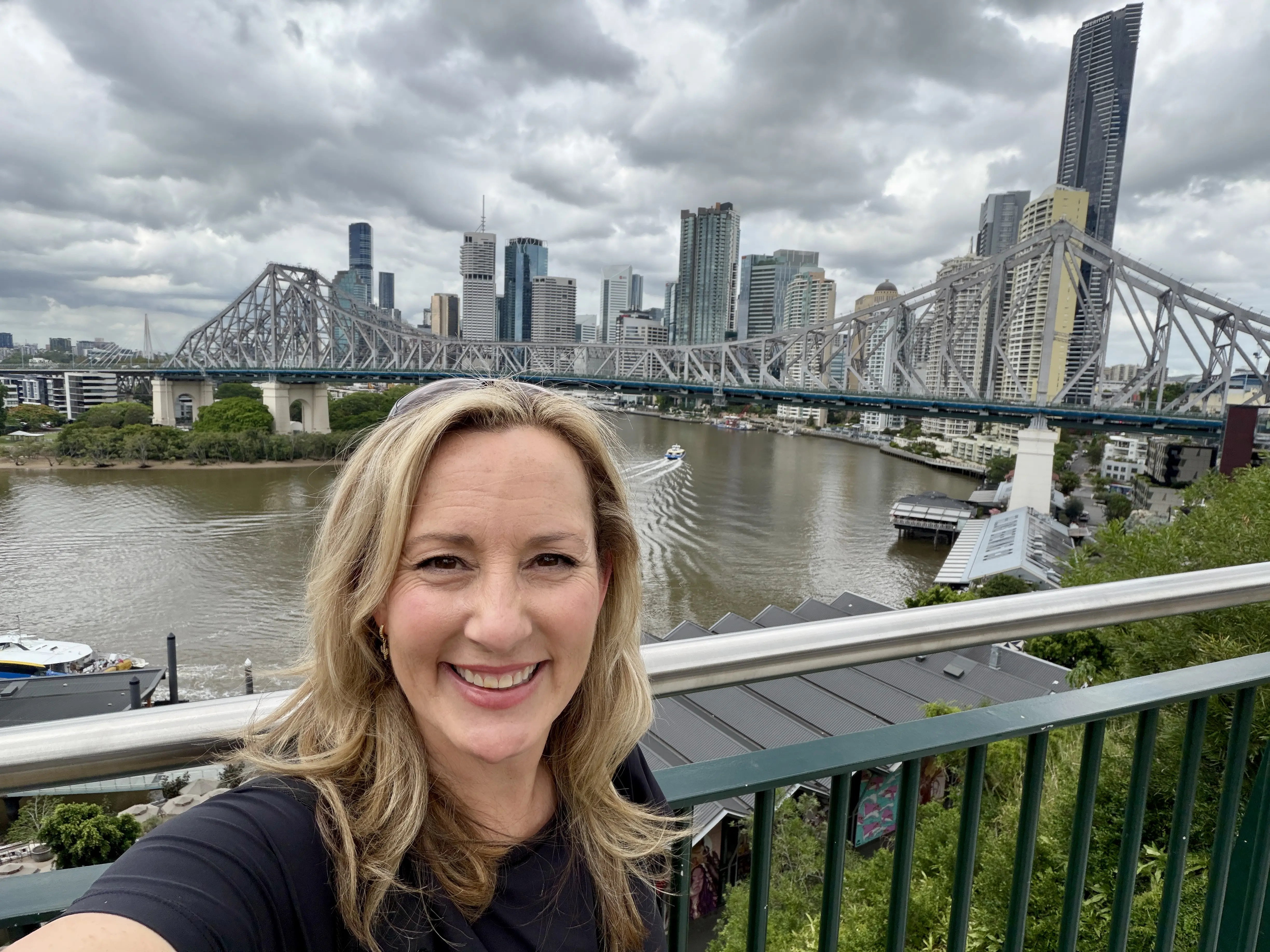 Blonde woman standing in front of bridge spanning a river. Skyscrapers in the background