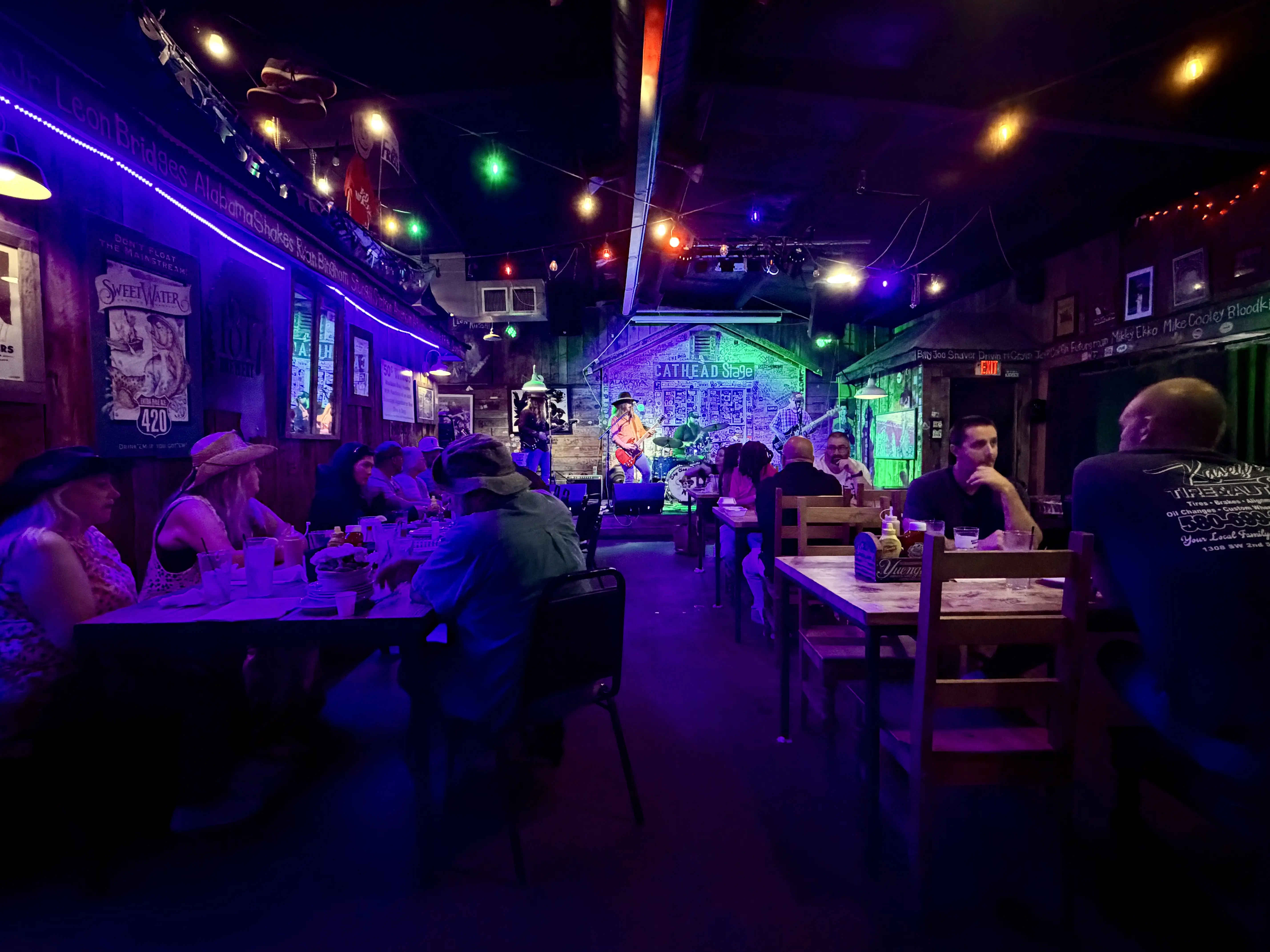 People sit at low top table in a dimly list bar. A blue led neon light lines the left wall giving the room a blue hue. At the back of the restaurant is a stage with a four man band playing guitars and drums.