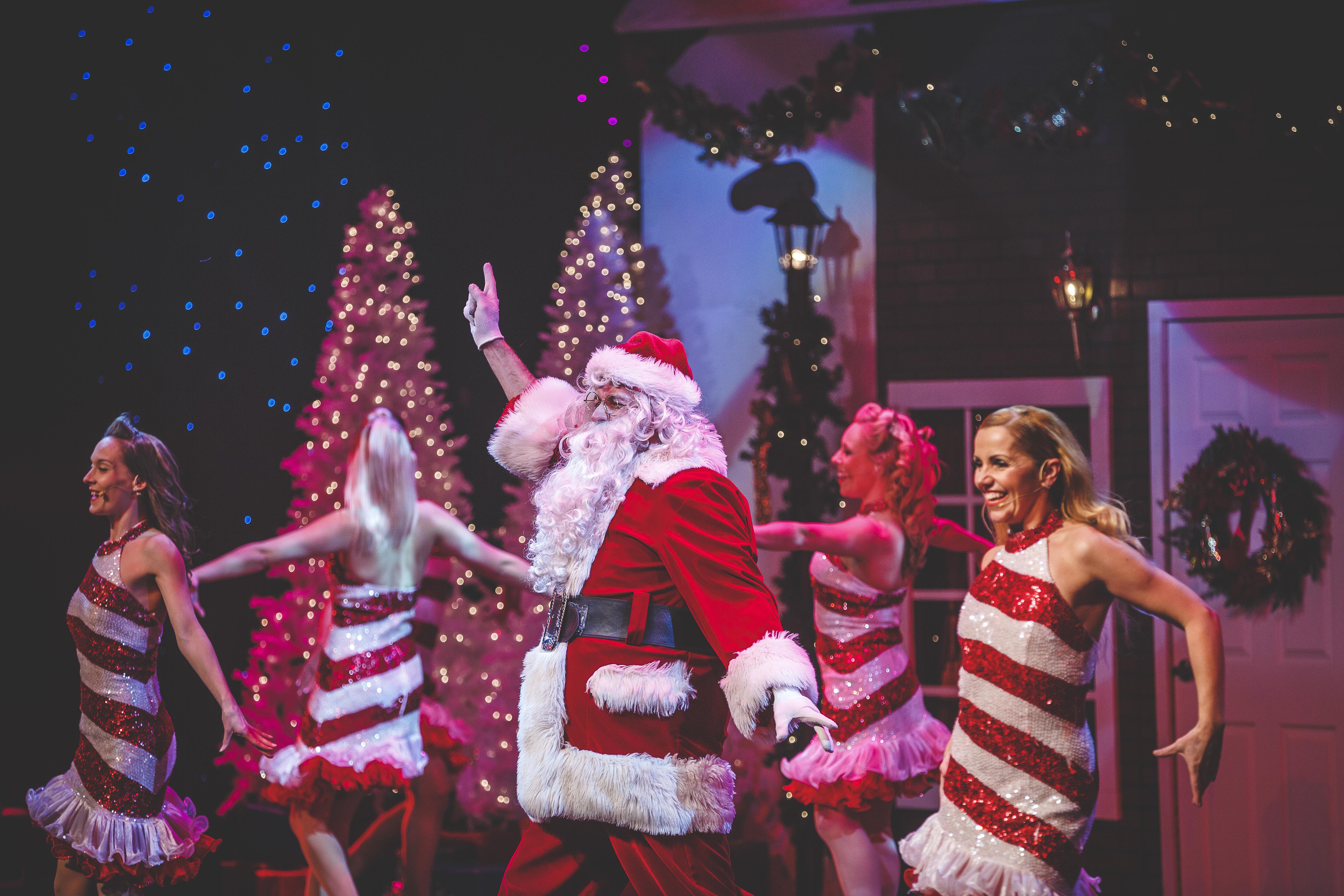 Man in red Santa costume performs on stage, accompanied by dancers in red and white candy cane striped dresses