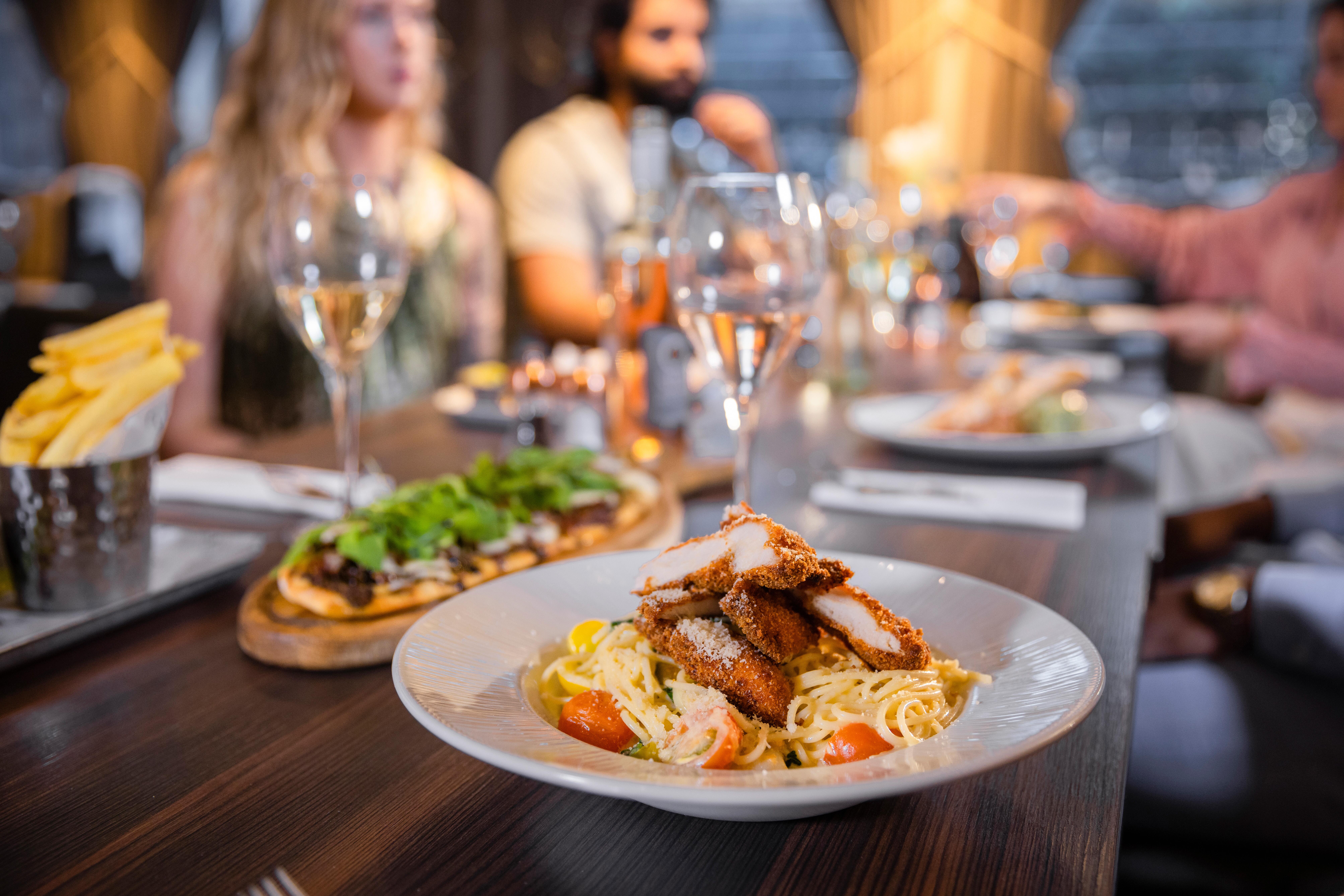 A close up shot of a bowl of pasta topped with breaded chicken on a table with a multi-ethnic group of friends wearing smart-casual clothing in a restaurant in the city on a winter's day