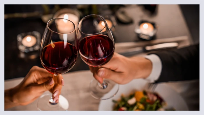 A closeup shot captures two hands holding glasses of red wine and clinking them together in a toast. 