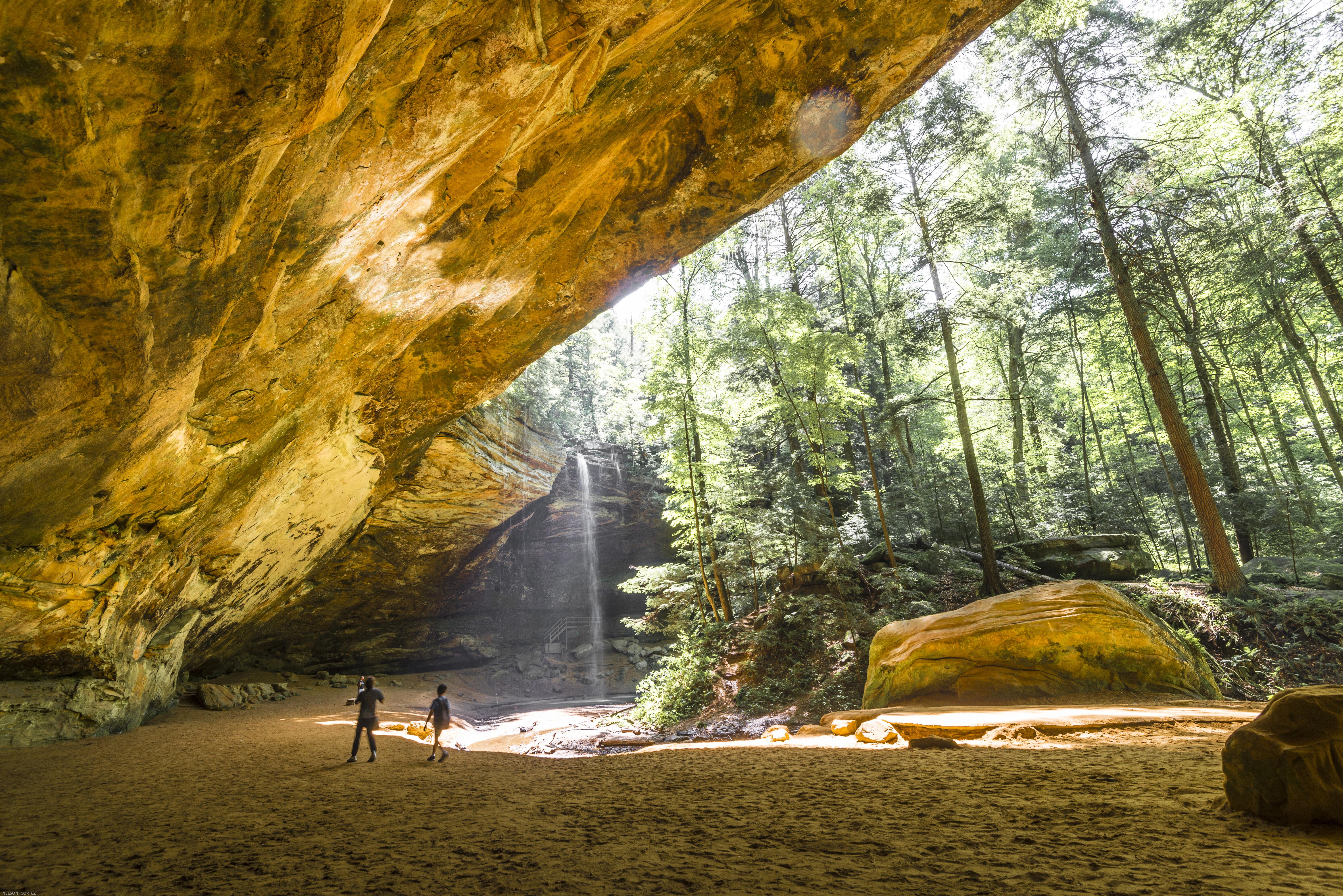 Two people in the distance under giant rock cave, amidst sunlit trees and waterfall in the forest of Hocking Hills State Park, Ohio