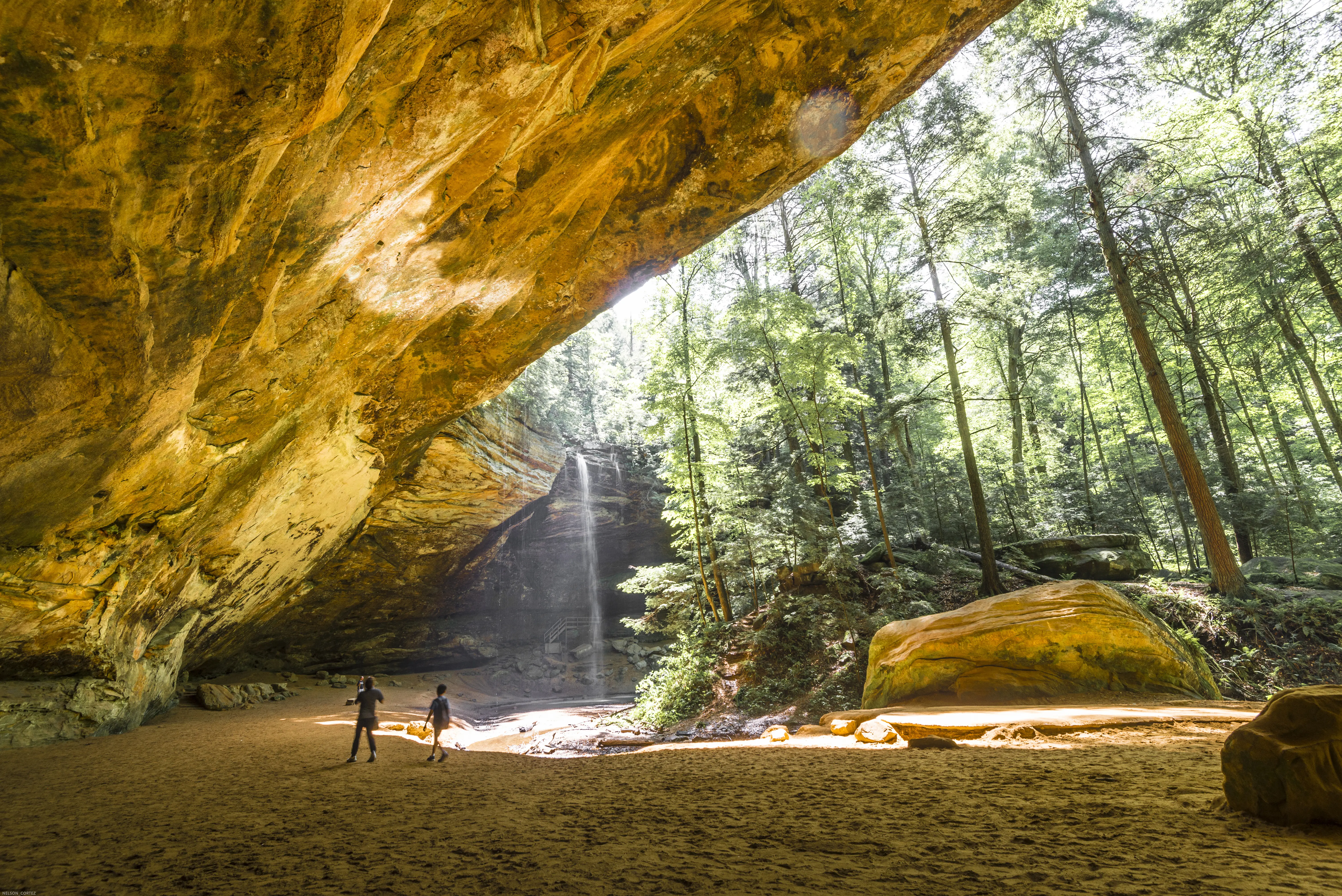 Two people in the distance under giant rock cave, amidst sunlit trees and waterfall in the forest of Hocking Hills State Park, Ohio