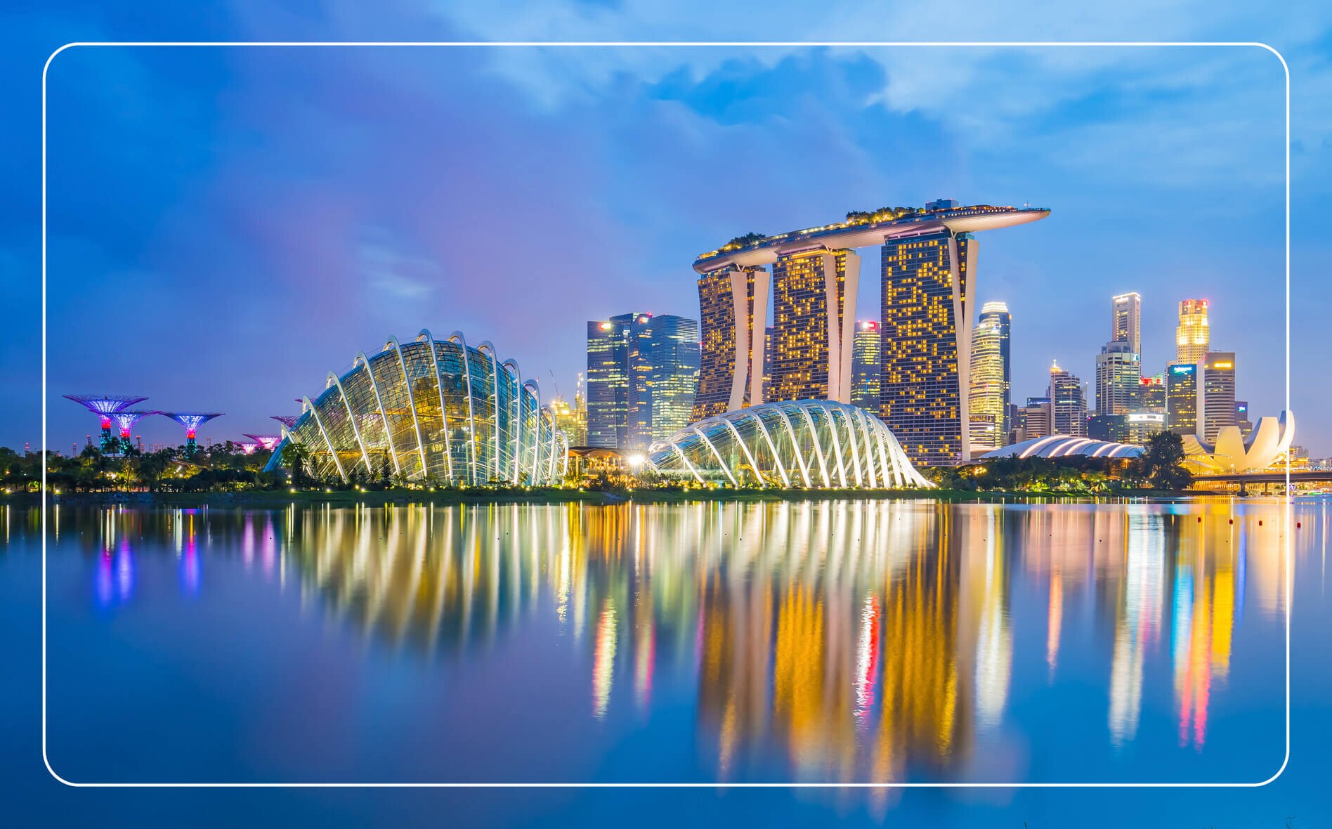 View of Singapore's skyline from the water at dusk, with dramatically illuminated modern skyscrapers rising against a blue and purple evening sky.