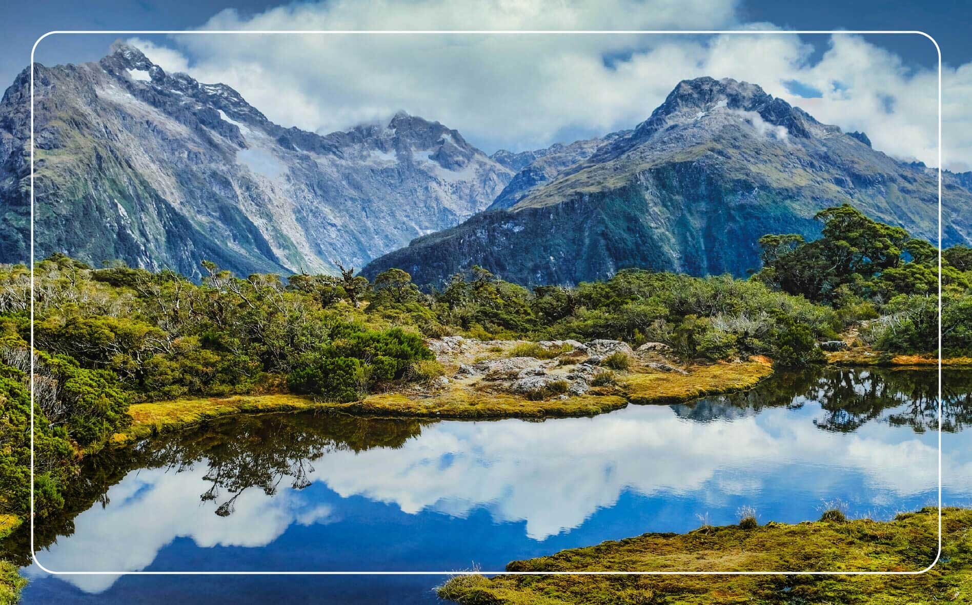 Aerial view of rugged mountains in New Zealand with fluffy clouds reflected in a tranquil mountain pond below.