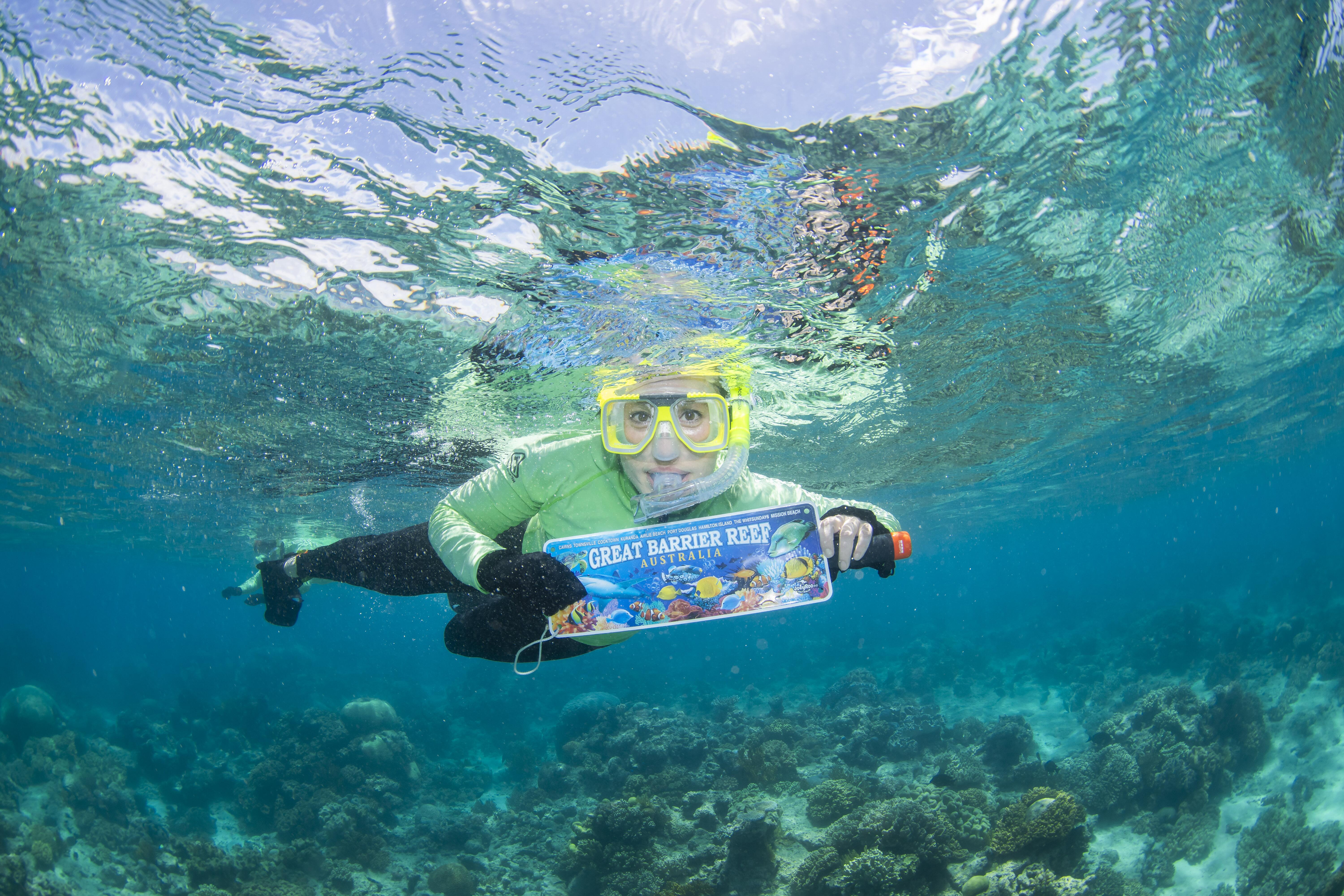 Woman underwater in snorkel gear holding small sign that reads Great Barrier Reef