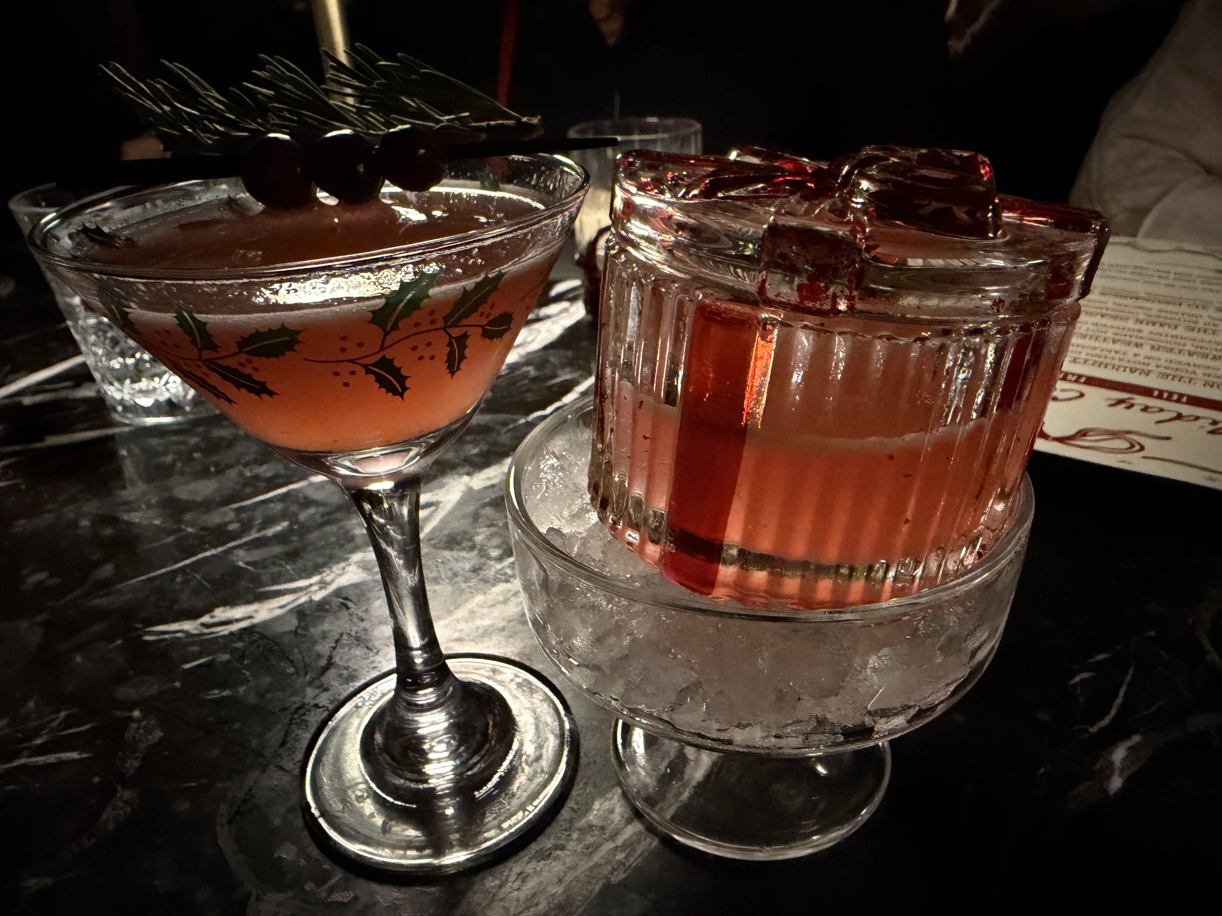 A smoky pink cocktail with a skewer of cherries and a sprig of thyme as garnish is served in a martini class with a festive holly decoration the encircles the glass.