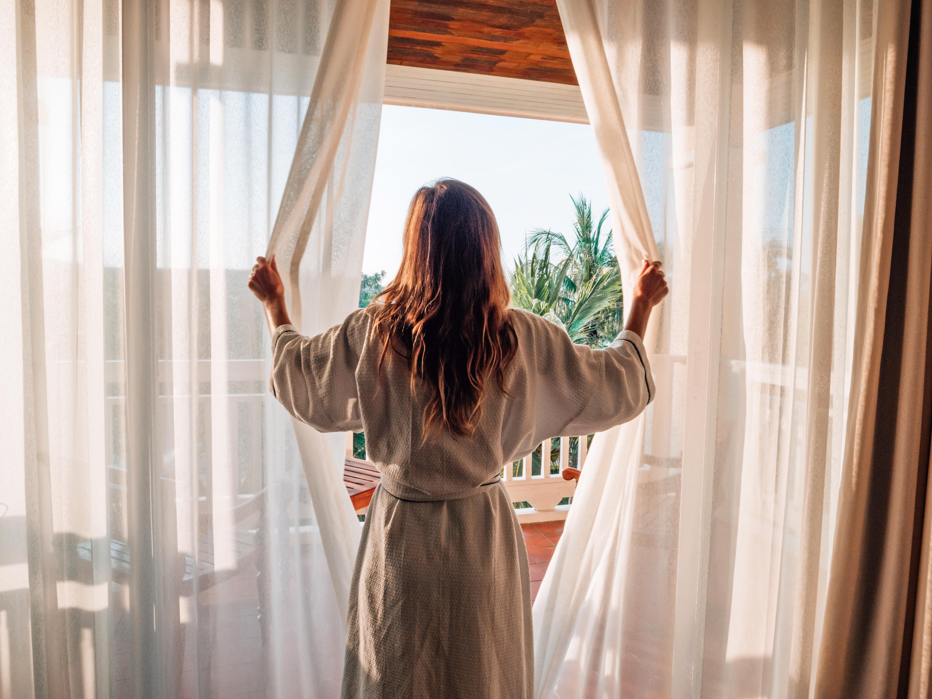Young woman in a white robe pulling open the curtains in a hotel room