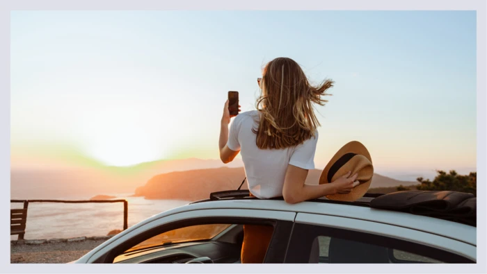 A woman is captured standing in the sky roof of her car while taking a photo of the beach shoreline and sunset. 